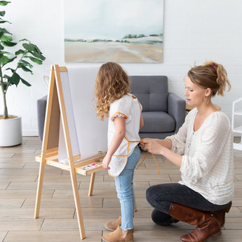 In a cozy room with wall art and plants, a woman adjusts a Bumkins Short-Sleeved Smock: Wander on a girl ready to paint at her easel.