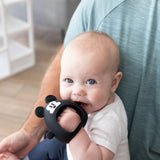 A blue-eyed baby nibbles on a Bumkins Silicone Un-Droppable Mitt Teether: Mickey Mouse while sitting on someone's lap indoors.