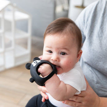 A baby in a white shirt chews on a Bumkins Silicone Un-Droppable Mitt Teether: Mickey Mouse while sitting on an adult's lap indoors.