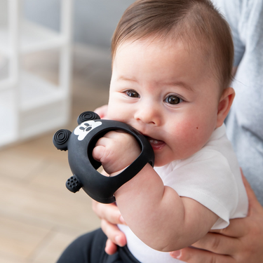 A baby in a white shirt chews on a Bumkins Silicone Un-Droppable Mitt Teether: Mickey Mouse while sitting on an adult's lap indoors.