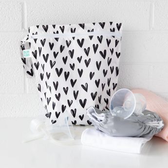 A Bumkins Wet Bag 2 Pack: Gray Arrow and Hearts sits on a white table against a white wall, with cloth diapers and breast pump parts in front.