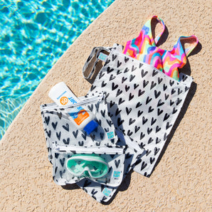 Bumkins The Chaos Coordinator Essential 4-Piece Set: Hearts holds a swimsuit, goggles, and sunscreen on a tan deck next to a blue pool.
