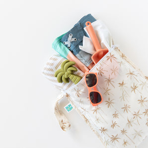 Bumkins Wet Bag + Wet/Dry Bag Set: Palm Daze, shown with towel, swimsuit, shovel & coral sunglasses on a white background.