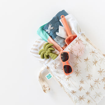 Bumkins Wet Bag + Wet/Dry Bag Set: Palm Daze, shown with towel, swimsuit, shovel & coral sunglasses on a white background.