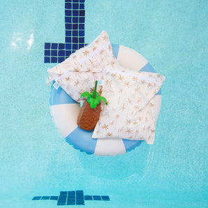 Bumkins Wet Bag + Wet/Dry Bag Set: Palm Daze rests with a palm tree shaped bottle atop a blue & white striped inner tube in a teal pool.