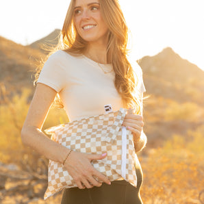 A woman carries a Bumkins Wet Bag: Sand Check in golden sunlight with mountains in the background.