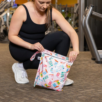 A woman in black athleisure wear kneels in a gym, unzipping a Roamio by Bumkins Wet Bag: Boots.