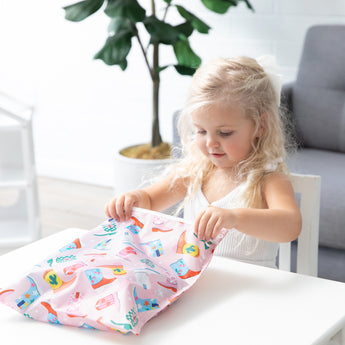 A blonde girl opens a bag from Bumkins Wet Bag + Wet/Dry Bag Set: Boots at a white table with a gray couch and plant behind.