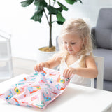 A blonde girl opens a bag from Bumkins Wet Bag + Wet/Dry Clutch Set: Boots at a white table in front of a plant and gray couch.