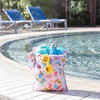 A bag from Bumkins The Chaos Coordinator Deluxe 5-Piece Set: Boots holds sunglasses and swim gear poolside with empty lounge chairs behind.