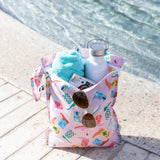 A bag from Bumkins The Chaos Coordinator Essential 4-Piece Set: Boots holds a towel, sunglasses, camera, and water bottle poolside.
