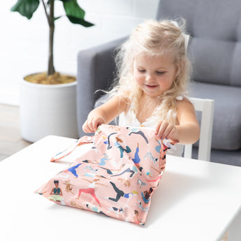 A blonde girl smiles as she opens a Bumkins Wet Bag: Yoga Pals at a white table in front of a gray couch and plant.