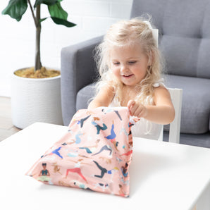 Smiling girl opens Bumkins Yoga Pals Wet Bag + Clear Travel Bag + Packable Tote Bag Set at a table.