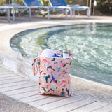 Bumkins Wet Bag: Yoga Pals holds a towel and water bottle poolside with empty lounge chairs in the background.