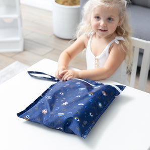 A smiling girl opens a bag from Bumkins Wet Bag + Wet/Dry Clutch Set: Evil Eye at a white table.