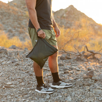 A man stands on rocky ground holding a Roamio by Bumkins Wet Bag: Rich Black with mountains in the background.