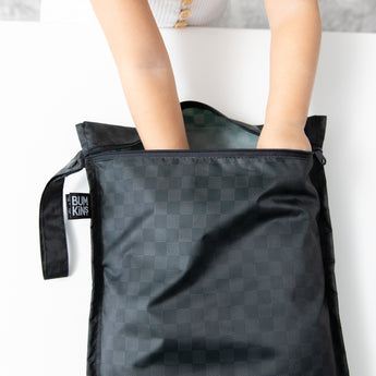 A child’s hands reach into a Bumkins Wet Bag 2 Pack in Sand and Black Check, made of waterproof fabric on a white surface.