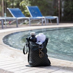 A bag from Bumkins The Chaos Coordinator Essential 4-Piece Set: Rich Black holds a towel, sunglasses, and water bottle poolside. 