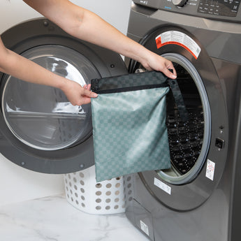 A person holds a bag from Roamio by Bumkins Wet Bag 2 Pack: Rich & Boujee in front of an open front-loading washing machine.