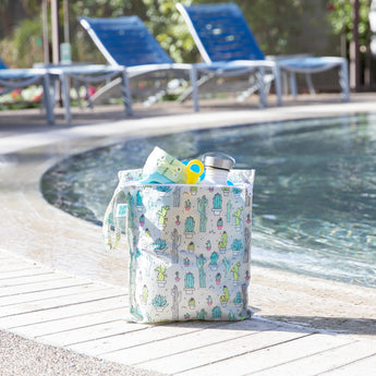 Bumkins Wet Bag: Cacti sits poolside, holding swimwear and a water bottle with empty lounge chairs in the background.