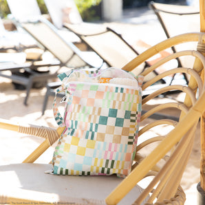 Roamio by Bumkins Wet Bag: Motley Check rests on a rattan chair poolside, holding a towel and sunscreen, with sun loungers nearby.