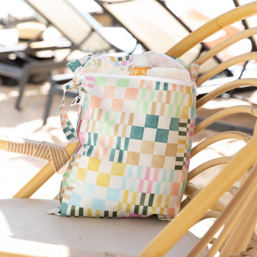 Roamio by Bumkins Wet Bag: Motley Check rests on a rattan chair poolside, holding a towel and sunscreen, with sun loungers nearby.