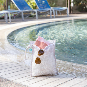 A bag from Bumkins Wet Bag 2 Pack: Wander and Sunshine holds sunglasses and swimwear poolside with empty lounge chairs in the background.