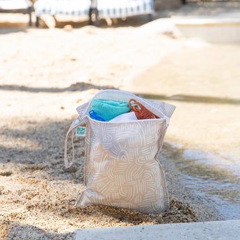 A bag from Bumkins Wet Bag + Wet/Dry Clutch Set: Wander sits on a sandy beach shore with a towel, swimwear, and goggles inside.