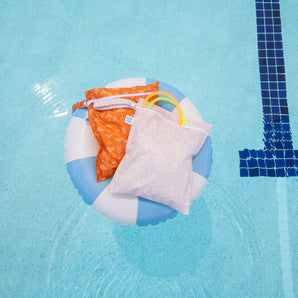 Bumkins Wet Bag 2 Pack: Wander + Grounded lays atop a blue & white striped inner tube in a pool; 2 diving rings peek out of the beige bag.