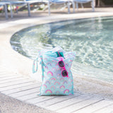 A blue Bumkins Wet Bag 2 Pack: Hearts and Rainbows holds sunglasses, a towel, and swimsuit poolside.