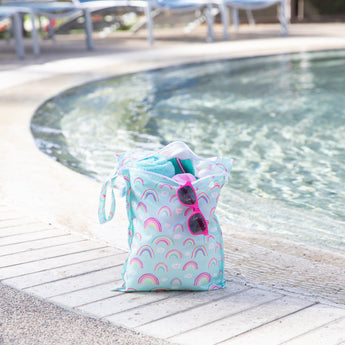 A blue Bumkins Wet Bag 2 Pack: Hearts and Rainbows holds sunglasses, a towel, and swimsuit poolside.