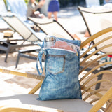 An open Roamio by Bumkins Wet Bag 2 Pack: Denim On Denim with a water bottle and pink cloth inside sits on a rattan lounge chair outdoors.