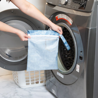A person holds a bag from Roamio by Bumkins The Commuter Essential 4-Piece Set: Denim on Denim in front of an open front-loading washing machine.