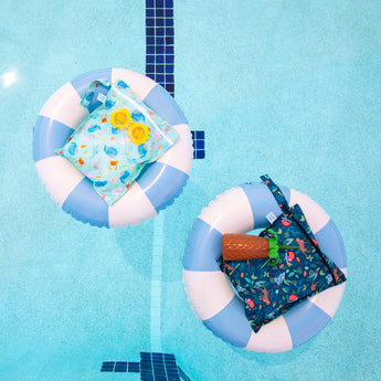 Bumkins Wet Bag 2 Pack: Ocean Life and Jungle lays in blue & white striped inner tubes with sunglasses & a palm tree shaped cup in a pool.