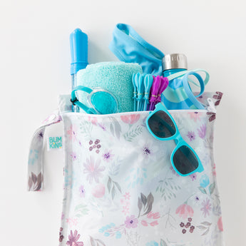 Bumkins Wet Bag: Floral holds a towel, goggles, sunglasses, swimsuit, and swim toys on a white background.
