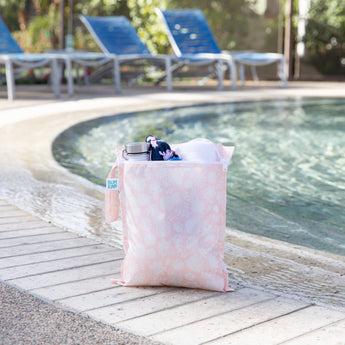 A bag from Bumkins The Chaos Coordinator Essential 4-Piece Set: Lace holds a water bottle, swimsuit, & towel poolside; lounge chairs behind.