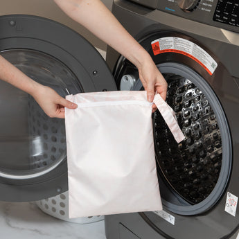 A person holds a bag from Bumkins The Chaos Coordinator Essential 4-Piece Set: Lace in front of an open front-loading washing machine.