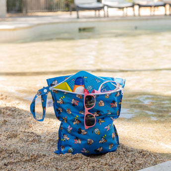 A bag from Bumkins Wet Bag + Wet/Dry Bag Set: Mickey & Friends Forever holds swim gear & sunglasses on a sandy beach.