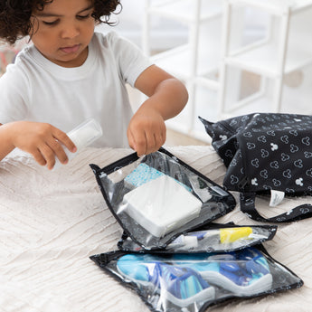 A young boy packs travel essentials in Bumkins The Chaos Coordinator Essential 4-Piece Set: Mickey Mouse Icon Black + White on a bed.
