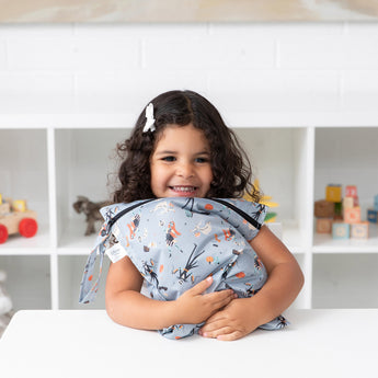 A smiling girl hugs a Bumkins Wet Bag 2 Pack: Everyday Is Halloween at a white table in front of toys on shelves.
