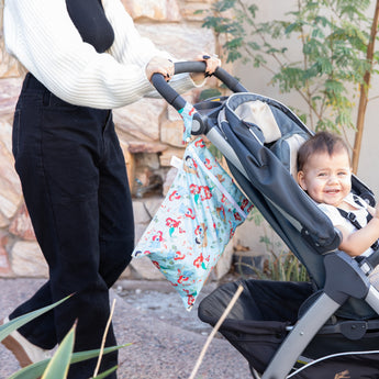 A person pushes a smiling baby in a stroller with a Bumkins Wet Bag: Disney Princess Ariel hanging from the handle.