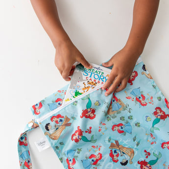 A child places a book into a Bumkins Wet Bag: Disney Princess Ariel, featuring The Little Mermaid, on a white table.