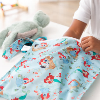 A child places items into a Bumkins Wet Bag: Disney Princess Ariel, with mermaid illustrations; a plush toy sits on the white table.
