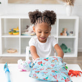 A smiling child with curly hair sits at a white table, reaching into a Bumkins Wet Bag: Disney Princess Ariel while surrounded by toys.