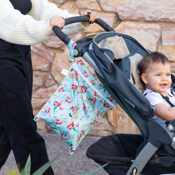 A person strolls with a baby in a stroller outdoors as a Bumkins Wet Bag: Disney Princess Ariel hangs from the handle.