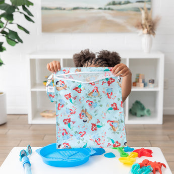 A child lifts a colorful Bumkins Wet Bag: Disney Princess Ariel at a white table with vibrant blue and red toys.