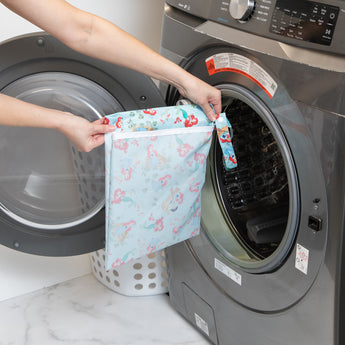 A person holds a Bumkins Wet Bag: Disney Princess Ariel in front of an open front-loading washer next to a white laundry basket.
