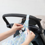 Hands attaching a Bumkins Wet Bag: Aloha Stitch strap to a black stroller handle, adding style and waterproof protection.
