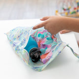 A hand reaches into Bumkins Wet Bag: Aloha Stitch, revealing a teal bottle and vibrant fabric on a pristine white table.