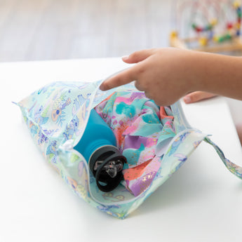 A hand reaches into Bumkins Wet Bag: Aloha Stitch, revealing a teal bottle and vibrant fabric on a pristine white table.